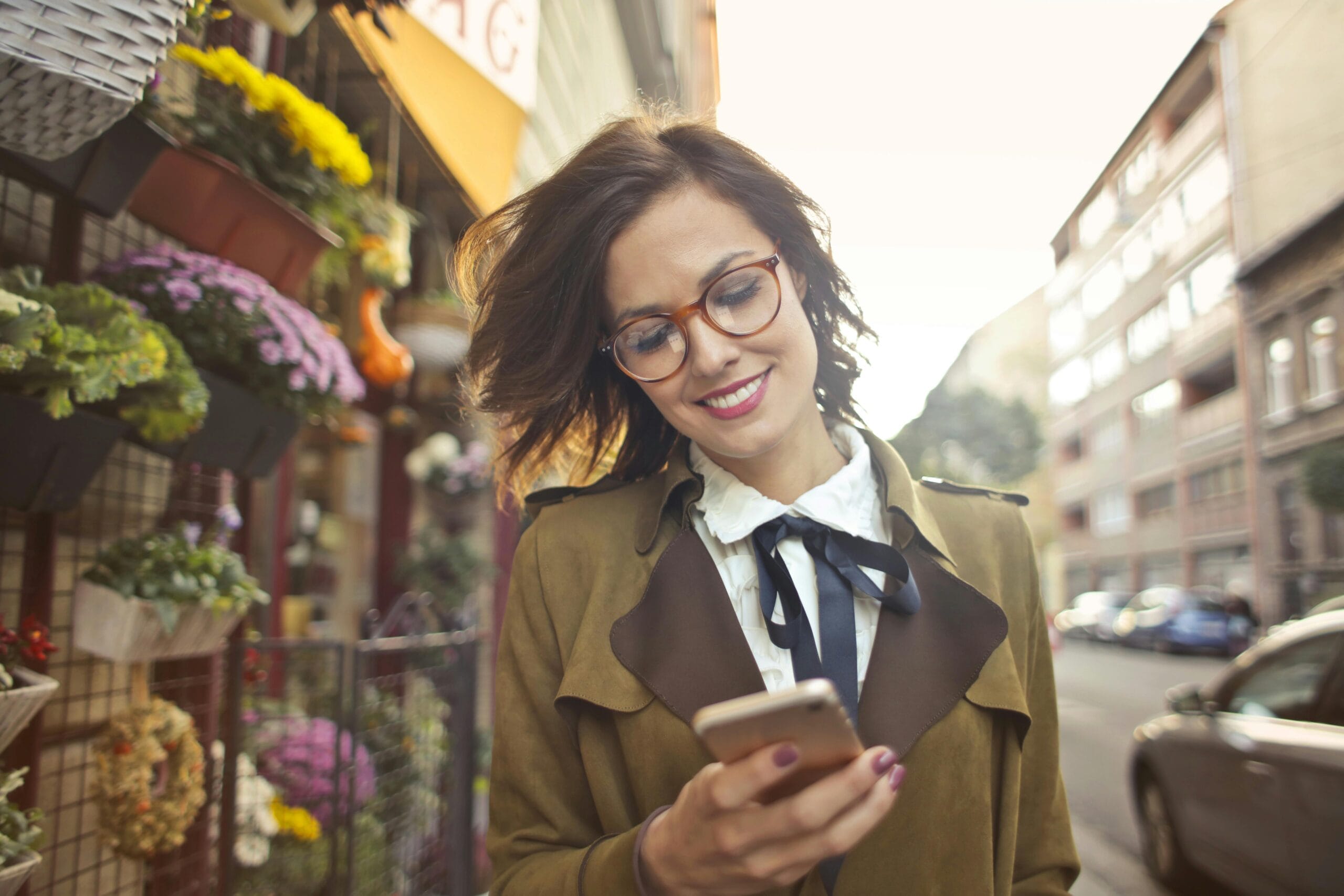 pexels-photo-3772531-3772531 A smiling woman uses her phone outside a colorful urban flower shop on a sunny day.