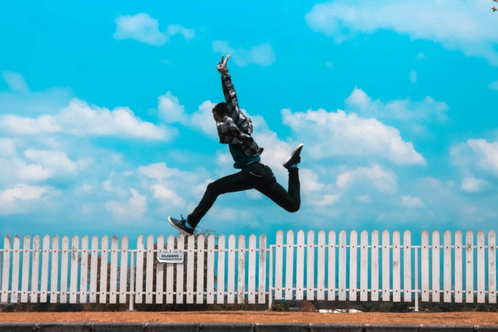 pexels-photo-1030794-1030794 Mid-air capture of a person leaping in front of a picket fence under a bright blue sky.